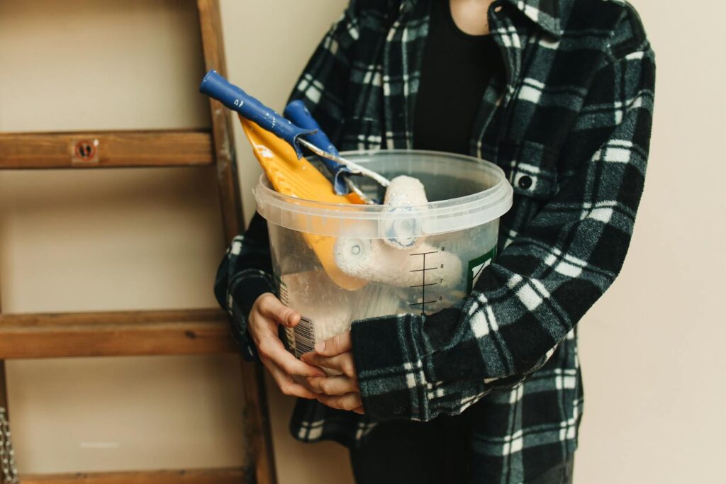 Close-up of a person holding a bucket filled with renovation tools, including paint rollers and scrapers.