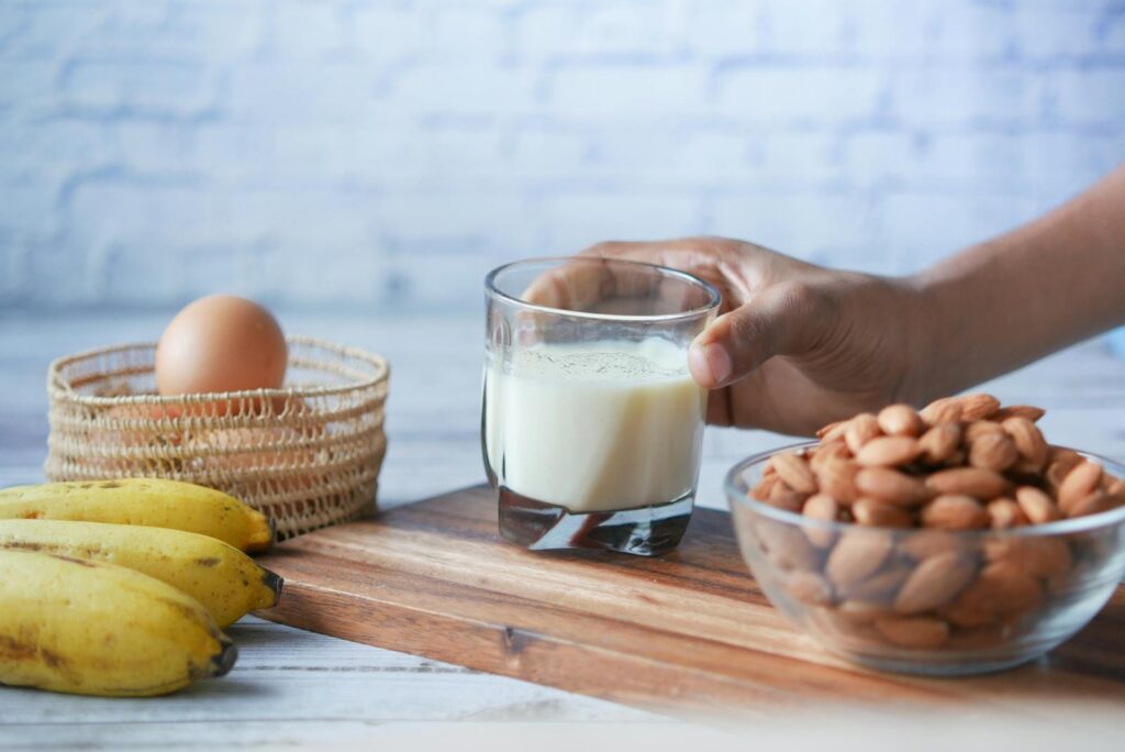 Close-up of a hand reaching for a glass of milk with almonds, banana, and egg on a wooden board.