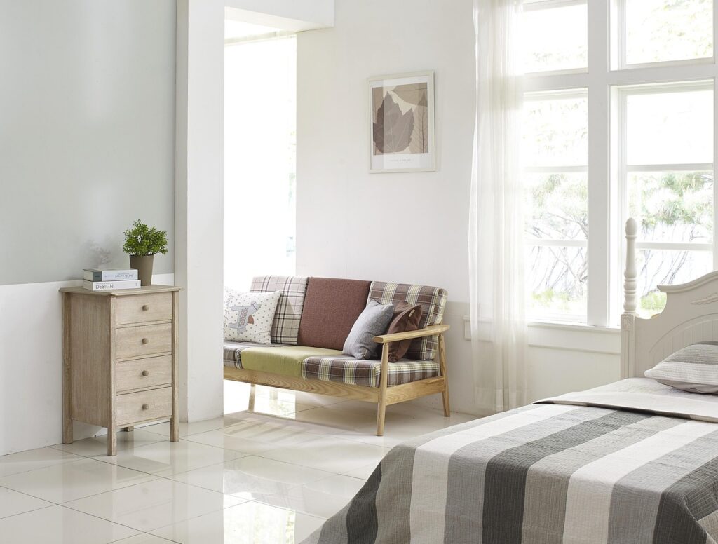 A minimalist white bedroom with a neatly arranged bed, a modern chair, a dresser, and a window allowing sunlight to enter.
