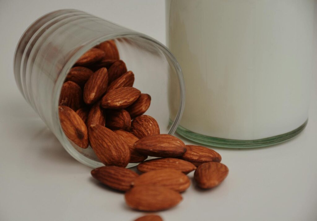 Raw almonds spilled from a glass next to almond milk on a white surface.