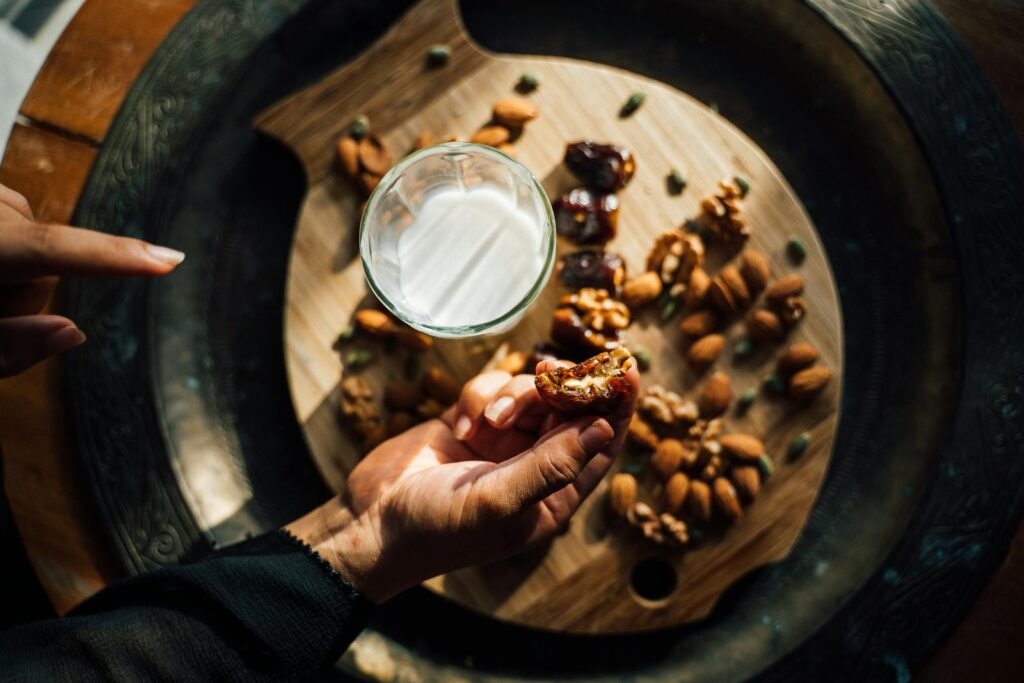 A hand holds a date near a wooden board with almonds, walnuts, and milk in natural light.