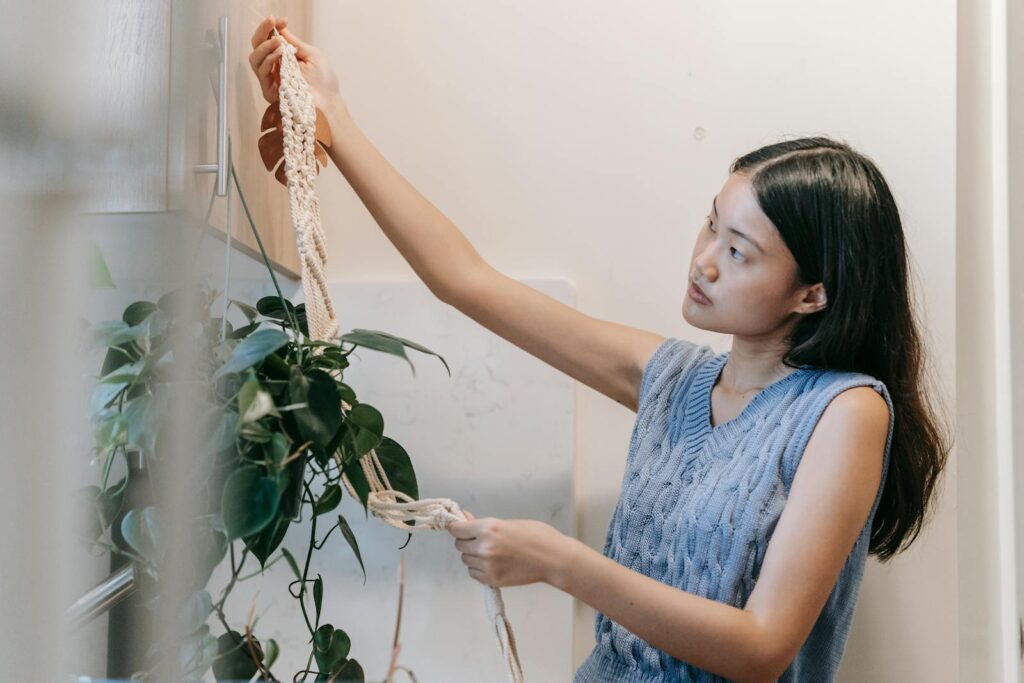 Young Asian woman arranging a macrame plant hanger in a cozy indoor setting.